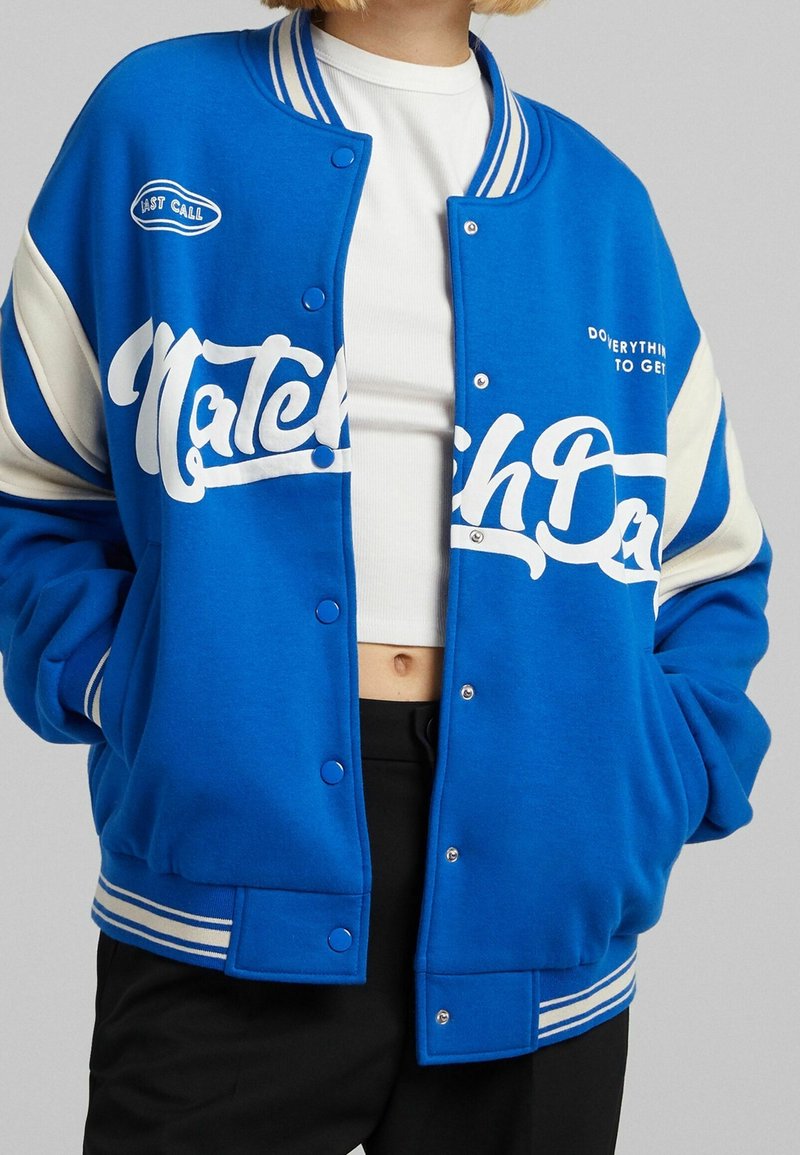 Niebieska kurtka varsity z białymi akcentami i napami, z napisem "Match Day" w pogrubionym, sportowym kroju na przodzie oraz prążkowanym kołnierzem.