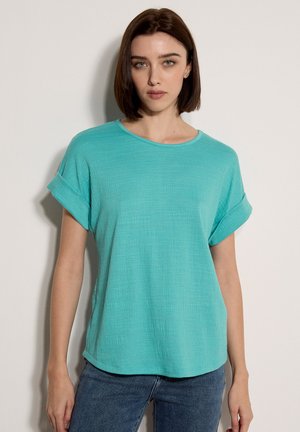 Donna con capelli castani lisci, che indossa una maglietta turquesa a maniche corte con texture e jeans blu, in piedi contro uno sfondo chiaro e uniforme.