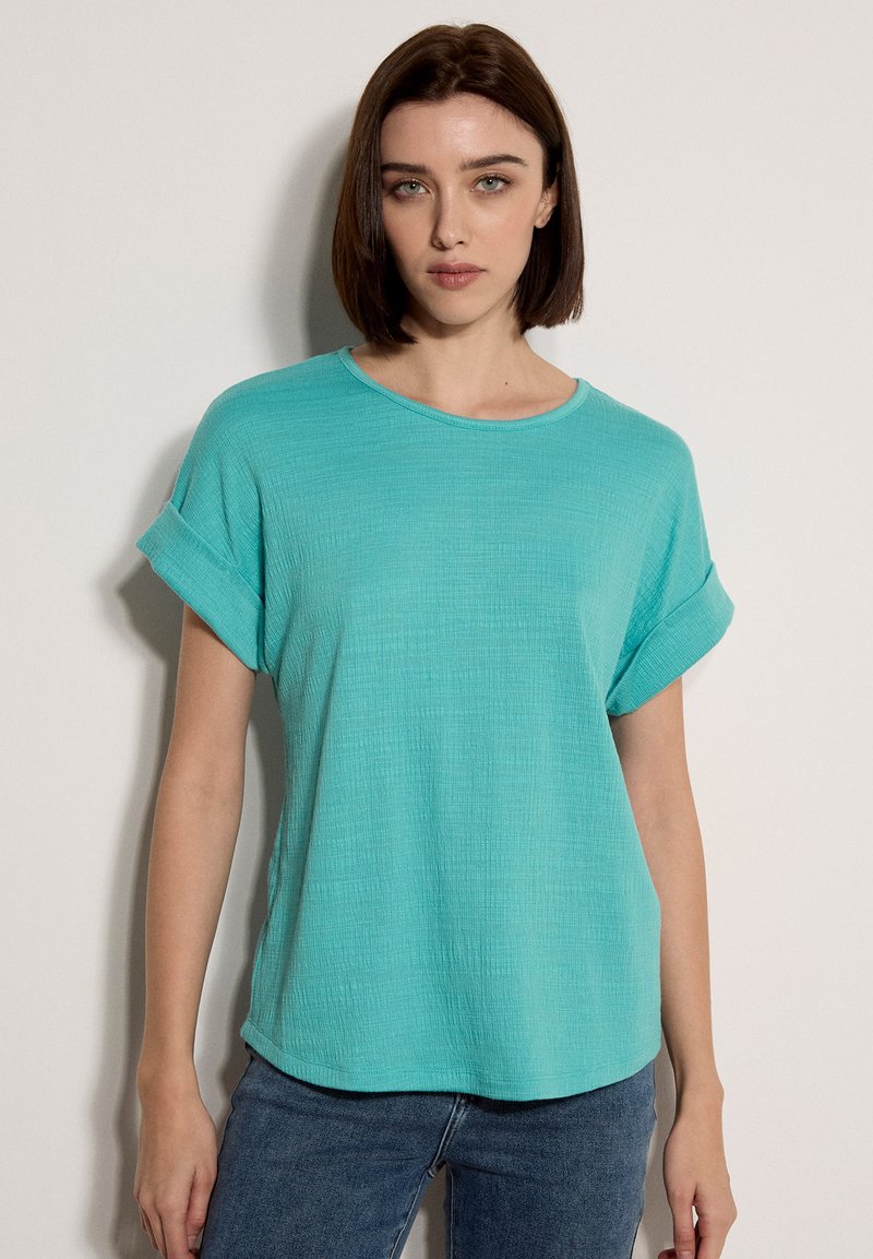 Femme aux cheveux bruns lisses portant un haut turquoise texturé à manches courtes et un jean bleu, debout devant un fond clair uni.