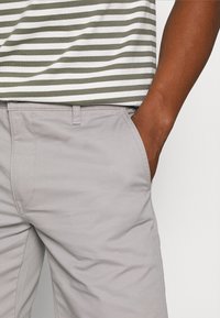 Personne portant un pantalon gris clair avec une main dans la poche et une chemise rayée verte et blanche rentrée.