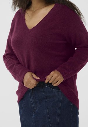 Femme portant un pull doux à manches longues, col en V bordeaux profond et un jean en denim foncé, ajustant l'ourlet du pull.