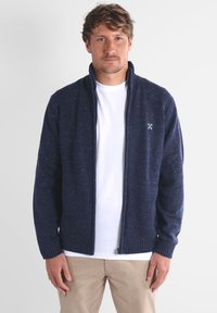 Gilet zippé marine en tissu tricoté texturé. Comprend des poignets et un ourlet côtelés. Porté par-dessus un t-shirt blanc, le modèle pose devant un fond uni.