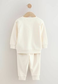Beige Baumwoll-Trainingsanzug auf einem Holzkleiderbügel aufgehängt. Beinhaltet ein Langarmoberteil und passende Hosen mit elastischen Bündchen.