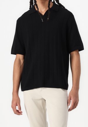 Czarna dzianinowa koszulka polo z krótkimi rękawami, z pionową prążkowaną fakturą i dwuknopkowym rozcięciem. W zestawie z jasnymi spodniami.