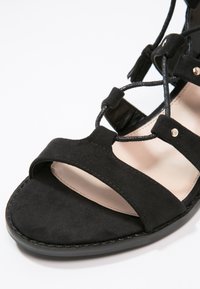 Sandale en daim noir avec un design croisé, bout ouvert et doublure intérieure lisse. Ornée de petits clous métalliques.