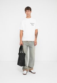 Witte korte mouwen t-shirt met zwarte tekst, lichtblauwe jeans en witte sneakers. Zwarte tote bag met textuur in de hand.