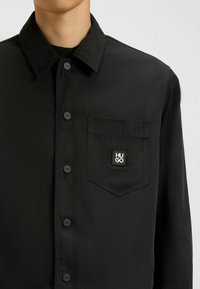 Musta takki, jossa on nappikiinnitys, terävä kaulus, rintatasku logopatchilla ja sileä, matta pinta.