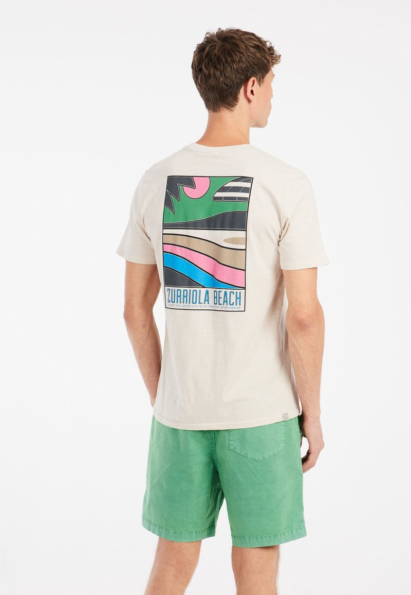 Kortærmet beige t-shirt med et farverigt grafik på ryggen, der viser bølger og palmetræer, med teksten "Zurriola Beach."