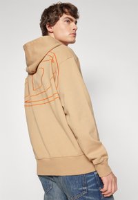Sudadera beige de corte holgado, con capucha y diseño de contorno naranja en la parte posterior. Fabricada con material suave y con puños acanalados.