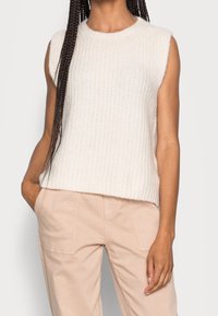 Haut sans manches en maille de couleur blanc cassé, avec une texture côtelée, un col rond et une coupe décontractée, porté avec un pantalon rose clair.