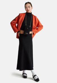 Junge Frau in einem schwarzen Rollkragen-Crop-Top, langer schwarzer Rock, orange Jacke, weißen Socken, schwarzen Schuhen und einer kleinen schwarzen Tasche mit goldener Kette.