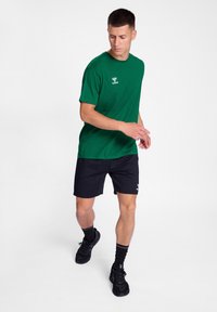 Groene katoenen T-shirt met een ronde hals en logodetail, gecombineerd met zwarte shorts. Model draagt zwarte sneakers en sokken. Witte achtergrond.
