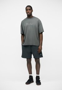 Noor mees, kes kannab suure suurusega halli T-särki tekstiga, musti lühikesi pükse, valgeid sokke ja musti kõrge säärega tossusid, seisab ühevärvilise tausta ees.