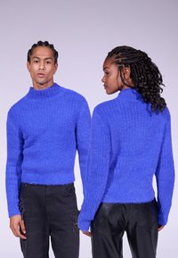 Pull bleu texturé à col montant avec un motif côtelé ; longueur cropped et manches longues, porté avec un pantalon sombre. Présente une finition douce et duveteuse.
