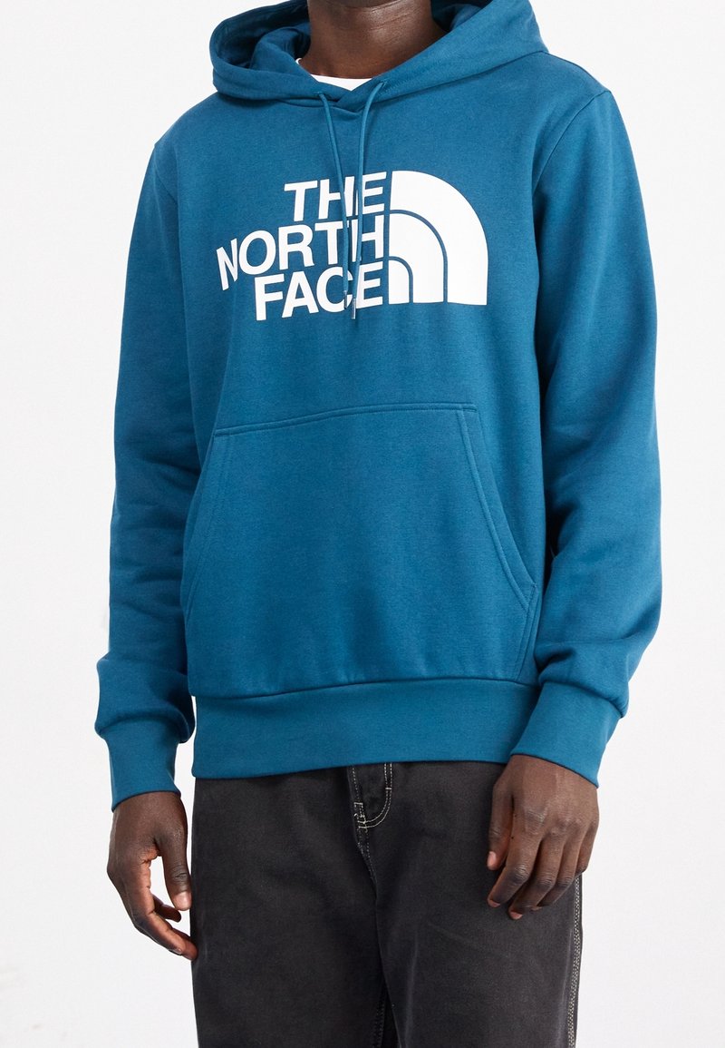 Personne portant un sweat à capuche bleu avec texte et logo « "THE NORTH FACE" » en blanc, assorti d'un pantalon noir.