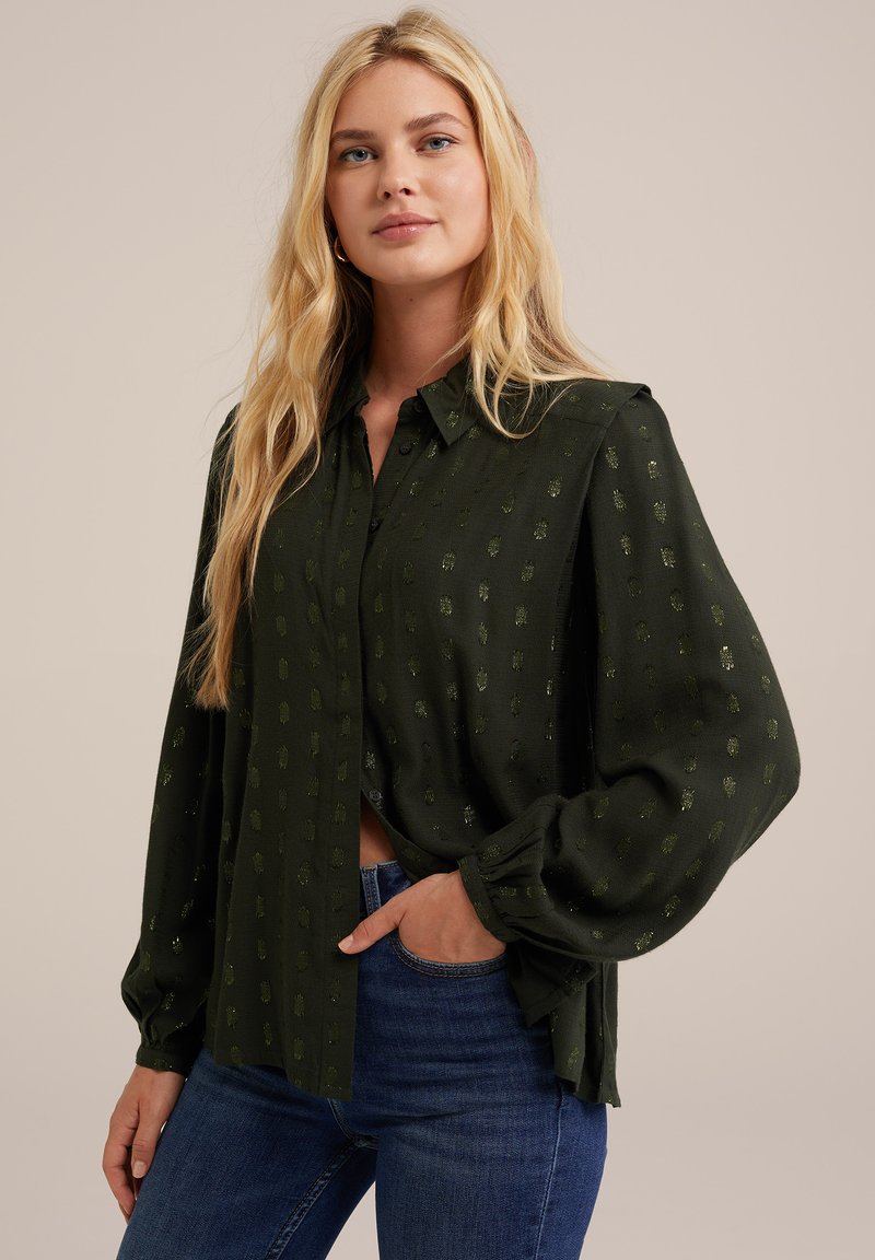 Blusa verde scuro con maniche lunghe a sbuffo e chiusura a bottoni. Presenta un motivo sottile e una consistenza morbida. Abbinata a jeans blu.