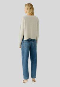 Hellbeige, verkürzter Pullover, lockere Passform, gerippter Saum. Hellblaue, hochgeschnittene Jeans, gerader Schnitt, dezente Details an den Gesäßtaschen, kombiniert mit hellen Absätzen.