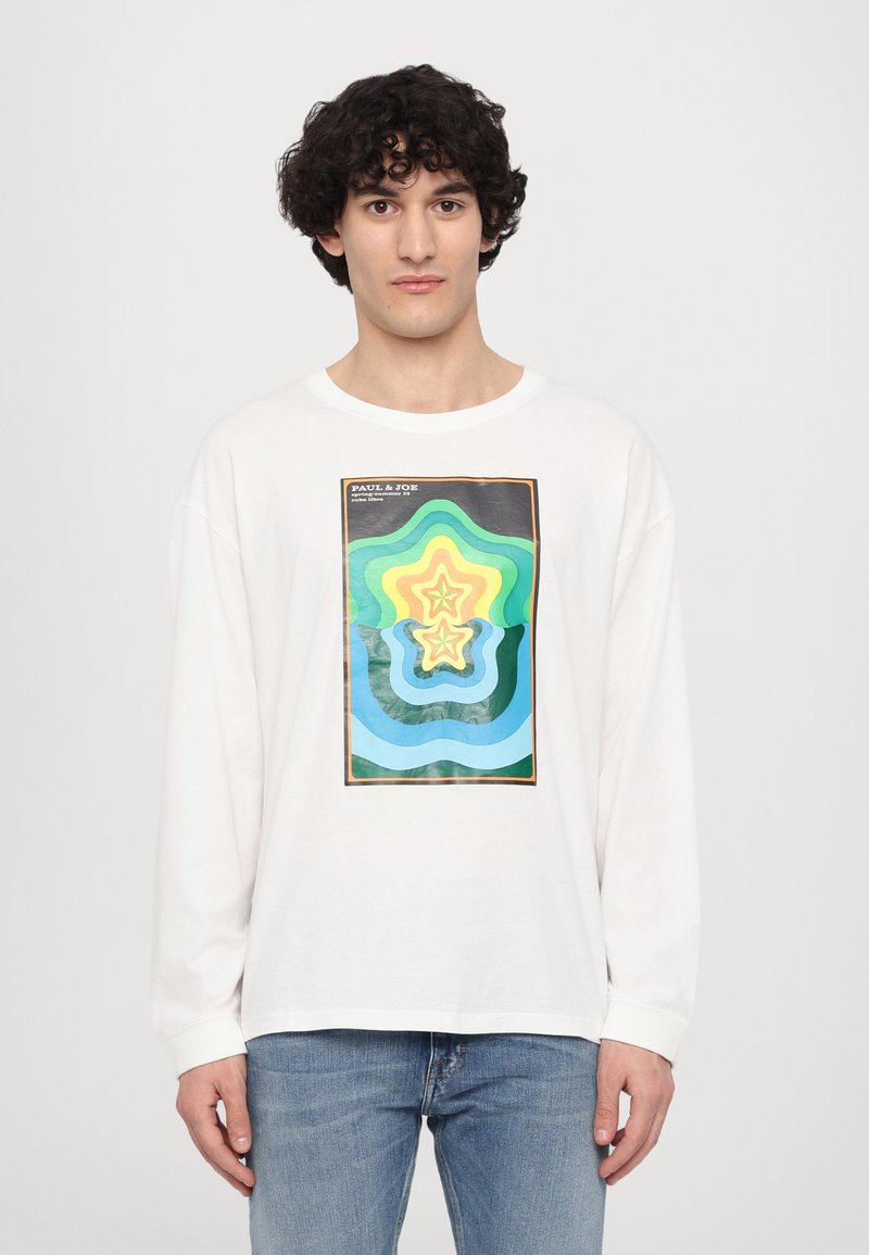 Witte lange mouwen t-shirt met een kleurrijke abstracte sterafbeelding in groen, blauw en geel, met een rechthoekige zwarte rand. Blauwe spijkerbroek.