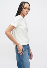 Lyse farget bomulls-t-skjorte med korte ermer, med rød tekst foran. Kombinert med høytlivs blå denimjeans.