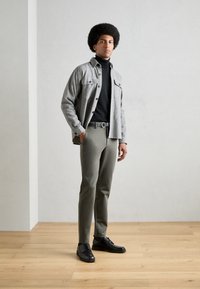 Chemise gris à boutons sur un col roulé noir, pantalon vert et chaussures noires. Tissu lisse au design épuré, se tenant devant un fond clair.