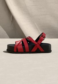 Sandale plateforme noire avec des lanières en cuir rouge croisées sur le pied et une fermeture à boucle argentée à la cheville.