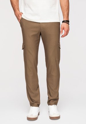 Man draagt bruine cargobroek met zijzakken, witte sneakers en een wit overhemd, staand tegen een effen achtergrond.