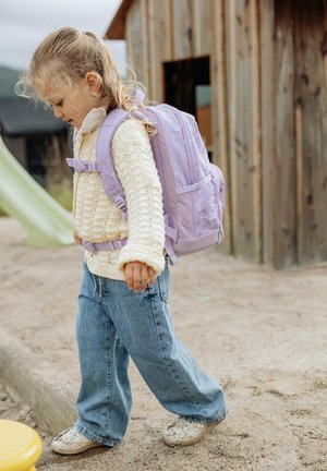 Ragazza giovane con zaino viola cammina su terreno sabbioso vicino a un edificio di legno e a uno scivolo del parco giochi, indossando un maglione e jeans.