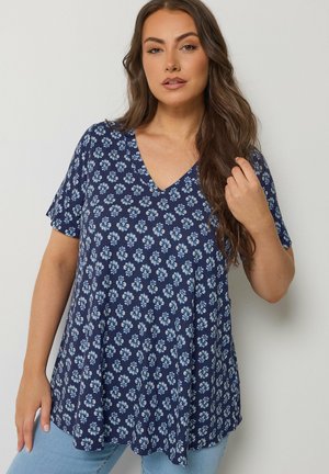 Kobieta z długimi brązowymi włosami, ubrana w niebieską kwiecistą bluzkę z krótkim rękawem i dekoltem w serek oraz jasnoniebieskie dżinsy na jednolitym tle.