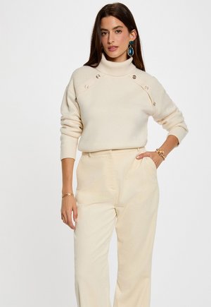 Maglione color crema con collo alto e texture a coste, dotato di bottoni decorativi sulle spalle, abbinato a pantaloni in corda a gamba larga dello stesso colore.