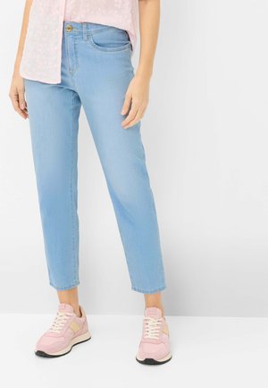 Person iført lyseblå jeans, pink blomsterprintet top og lyserøde sneakers, stående mod en ens hvid baggrund.