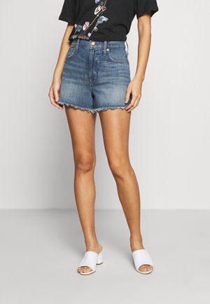Kvinde iført slidte denimshorts med rå kant, sort T-shirt med blomsterprint og hvide blokhælede sandaler med åben tå, stående mod en grå baggrund.