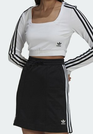 Kobieta nosząca biały krótki top z długim rękawem i czarną spódnicę, oba z logo Adidas oraz trzema czarnymi lub białymi paskami na rękawach i bokach.