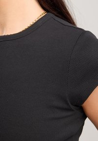Black ribbed short-sleeve top featuring a round neckline, soft texture, and a fitted design, complemented by a thin gold chain necklace.