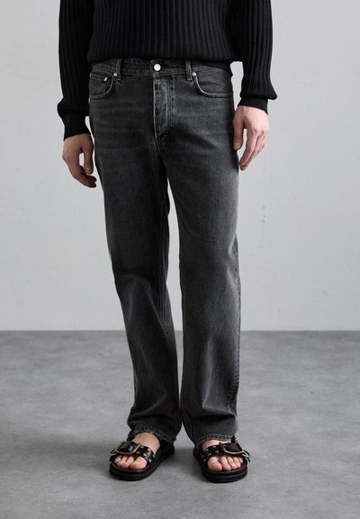 Sort ribbet striktrøje, grå jeans med lige ben, og sorte sandaler med hardware-accenter. Glat stof og minimalistisk design.