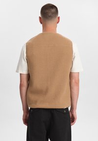 Gilet di feltro beige con colletto arrotondato; taglio dritto; vista posteriore senza chiusure; indossato sopra una camicia bianca a maniche corte; pantaloni neri.