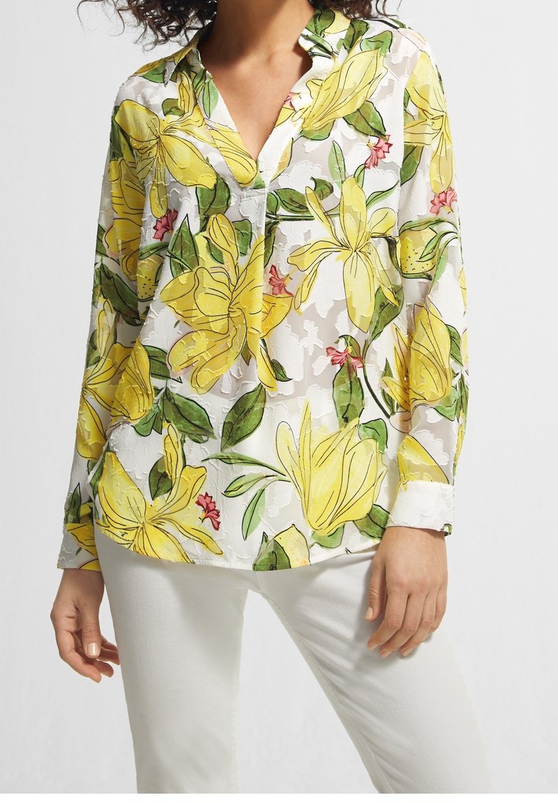 Blouse à manches longues florale en tissu blanc transparent, ornée de fleurs jaunes vives et de feuilles vertes, avec un col en V et une coupe décontractée.