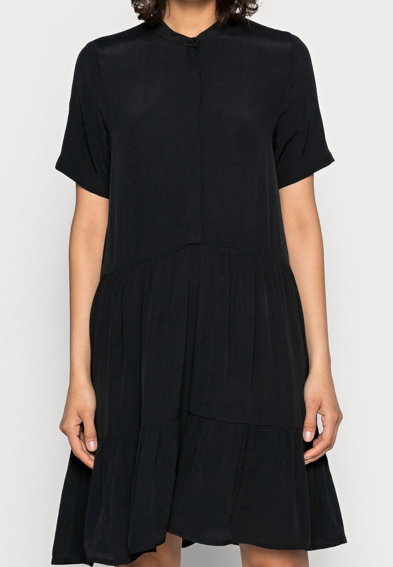 Femme portant une robe noire à manches courtes avec un col montant et une jupe plissée, debout devant un fond uni.
