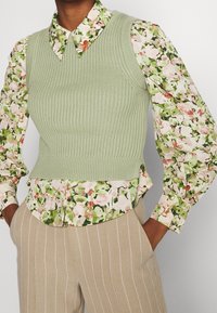 Světlá zelená vestička z žebrovaného úpletu přes květinovou halenku se zelenými a růžovými akcenty. Má límec a široké rukávy.