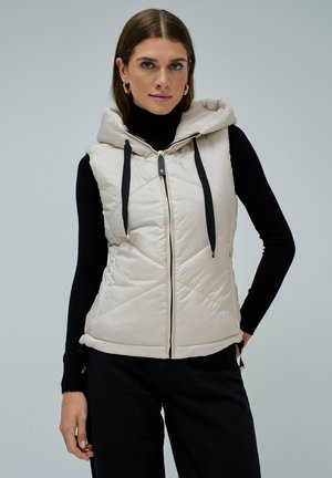 Donna che indossa un gilet imbottito beige con cappuccio e zip nera sopra una maglia nera a maniche lunghe e pantaloni neri, in piedi davanti a uno sfondo semplice.