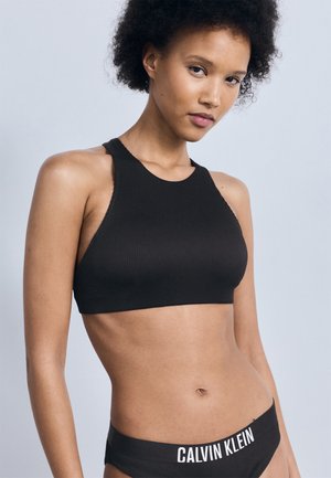 Donna con capelli ricci corti che indossa un top nero senza maniche e mutande nere Calvin Klein su uno sfondo grigio chiaro.