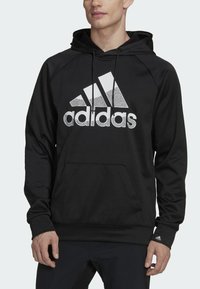 Felpa nera Adidas con un grande logo bianco. Presenta un cappuccio con stringa e tasca a marsupio. Realizzata in tessuto liscio e leggero.
