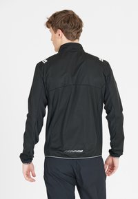 Svart idrottsjacka med hög krage, reflexdetaljer på axlarna och en texturerad bakpanel. Lätt och åtsittande design.