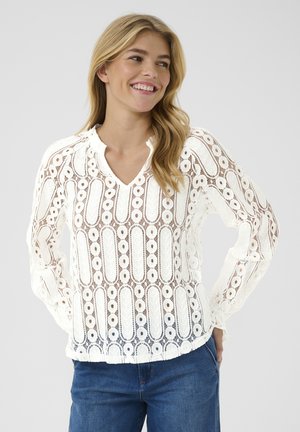 Weißes Spitzenbluse mit V-Ausschnitt, langen Ärmeln und aufwändigen gemusterten Ausschnitten. Kombiniert mit blauen Jeans, die ein leichtes, strukturiertes Design zeigen.
