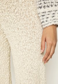 Cremfarbene gestrickte Hose mit einem strukturierten Muster, die eine lockere Passform hat, und eine Nahaufnahme einer Hand, die sanft den Stoff berührt.