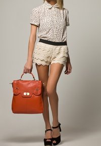 Orange läderryggsäck med en överdel, guldiga detaljer och dubbla handtag. Bärs med en prickig skjorta och beige vågiga shorts. Svarta klackskor.