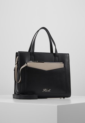 Sac à main en cuir noir avec double poignée, poche avant contenant une pochette beige zippée et logo argenté sur une surface blanche sur fond gris.