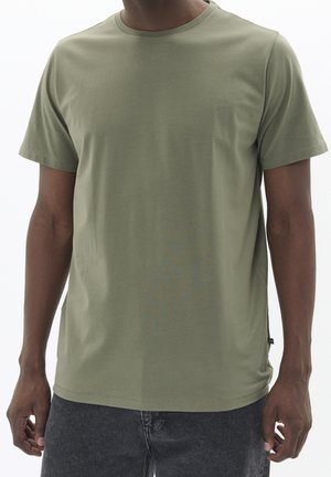 Man die een effen olijfgroen T-shirt met korte mouwen en donkergrijze spijkerbroek draagt, staand tegen een effen lichte achtergrond.
