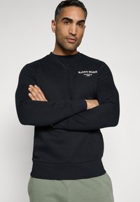 Črna sweatshirt s krožnim izrezom in dolgimi rokavi, z belim logotipom na prsih. Gladek material z oprijetim dizajnom.