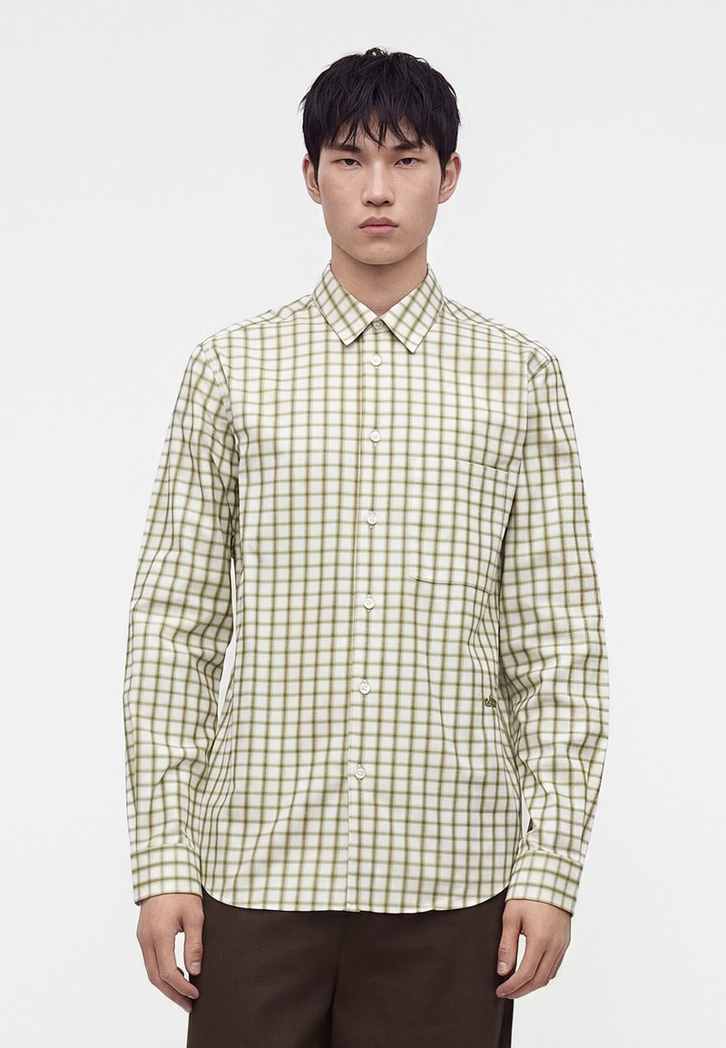 Homme portant une chemise blanche boutonnée avec un motif à carreaux verts et un pantalon marron foncé sur un fond blanc uni.