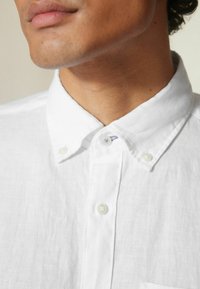Hvid, knap-op skjorte lavet af hør; har en blød tekstur, spidse kraver og synlige knapper med et lille accent af lilla syning.